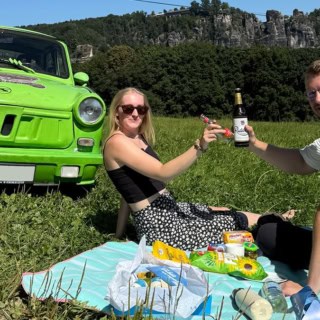 Foto von einem grünen Trabant mit Mann und Frau davor, auf einer Picknick decke sitzend beim Picknick. Im Hintergrund die Sächsische Schweiz