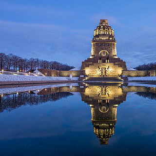 Beleuchtetes Völkerschlachtdenkmal spiegelt sich im ruhigen Wasser, verschneite Ufer und blaue Abendstimmung.