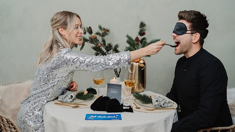 Paar füttert sich mit dem Inhalten der Geschenkbox Dinner in the Dark