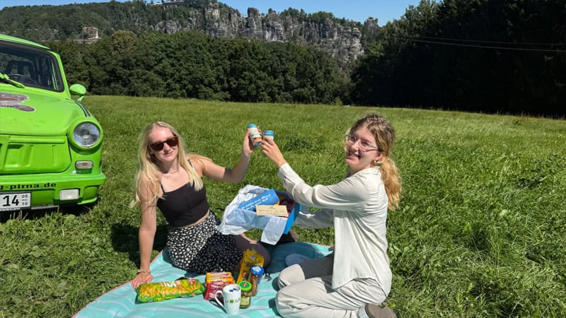 Zwei Frauen auf der Wiese beim Trabi Picknick packen die Geschenkbox für Trabant Erlebnisse aus