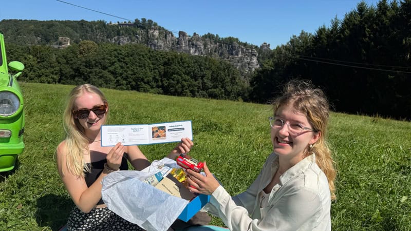 Zwei Frauen sitzen auf der Wiese in der Sächsischen Schweiz und halten die Gutscheine hoch, die in der Geschenkbox der Trabi Erlebnisse enthalten sind.