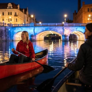 zwei Frauen paddeln in Kanus bei Dämmerung unter einer beleuchteten Brücke in Leipzig