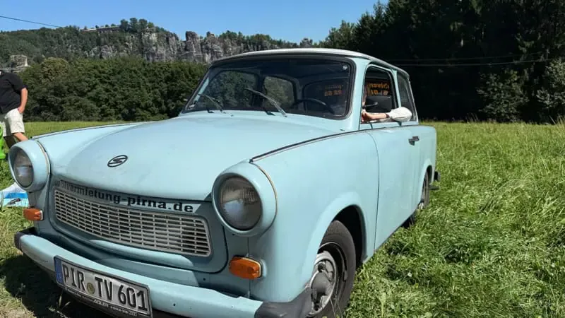 Blauer Trabant aus DDR-Zeiten steht allein auf einer grünen Wiese vor steilen Sandsteinfelsen im Sonnenschein.