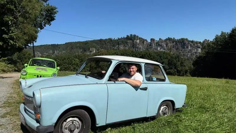 Mann lehnt sich aus dem Fenster eines blauen Trabant, hinten fährt ein grüner Trabant vorbei, Felswände im Hintergrund.