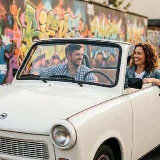 ein Paar fährt lächelnd in einem weißen Trabant Cabrio durch Chemnitz vor einer Graffiti-Wand