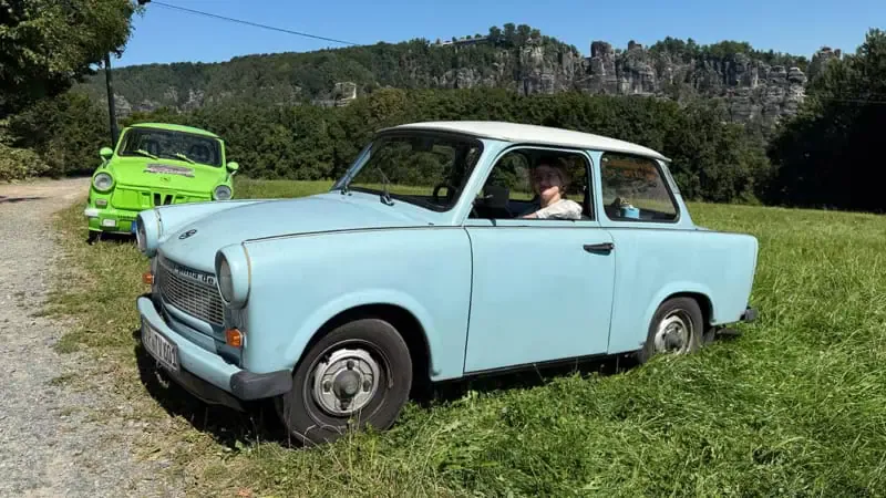 Blauer Trabant fährt über eine Wiese, ein grüner Trabant folgt im Hintergrund, dahinter Sandsteinfelsen und Wälder.