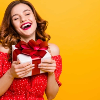 Eine Frau in einem roten Kleid hält lachend ein Geschenk in einer weißen Verpackung mit einem roten Geschenkband.