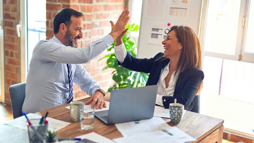 Zwei Personen sitzen am Tisch, arbeiten und sind glücklich. Sie geben sich dabei gegenseitig ein High-Five. Im Hintergrund Flipcharts mit Diagrammen, welche Erfolge darstellen sollen.