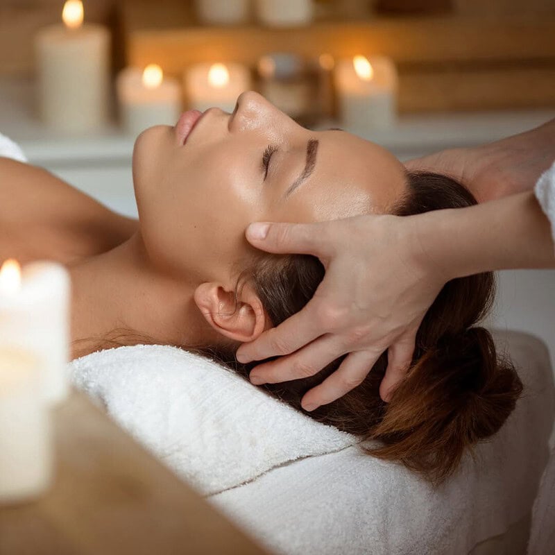 Frau liegt entspannt auf einer Liege und bekommt eine sanfte Kopfmassage, Kerzen im Hintergrund schaffen Wohlfühlatmosphäre.