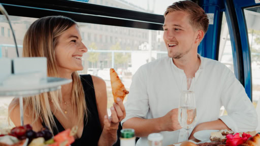 Frau und Mann sitzen in dem riesenrad in Dresden und frühstücken. Mann rechts hat ein weißes Hemd und die Frau ein schwarzes Oberteil an.