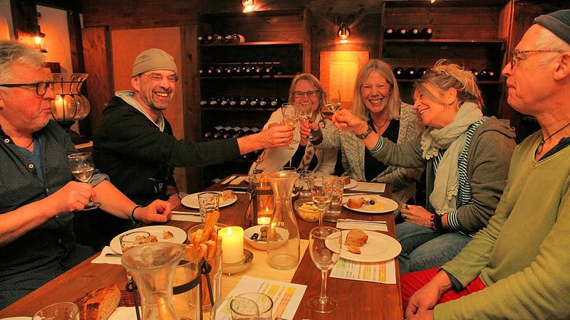 3 ältere Paare stoßen an einem mit Beilagen gedeckten Tisch an. Sie befinden sich in einem gemütlichem Wein Restaurant.