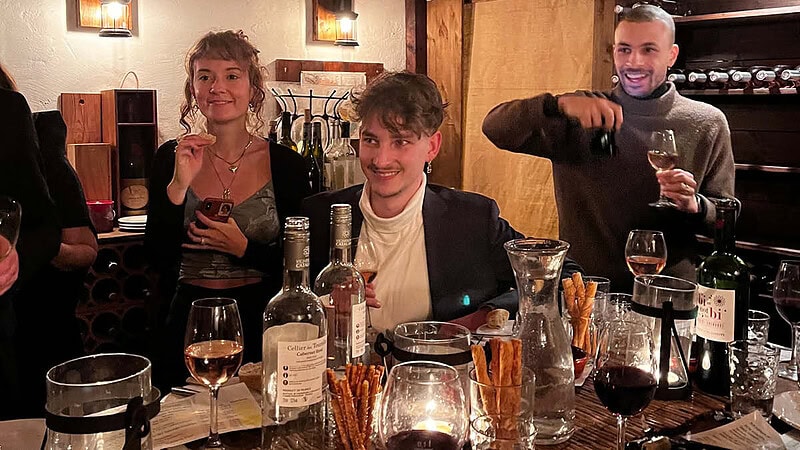 3 Leute die an einem mit Wein und Beilagen gedecktem Tisch sitzen und Lachen.