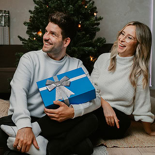 Mann und Frau mit der Geschenkbox der Erlebnisfabrik in der Hand schauen nach links, im Hintergrund befindet sich ein Weihnachtsbaum