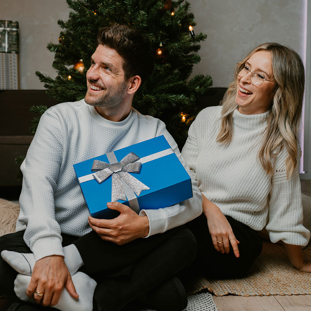 Mann und Frau mit der Geschenkbox der Erlebnisfabrik in der Hand schauen nach links, im Hintergrund befindet sich ein Weihnachtsbaum