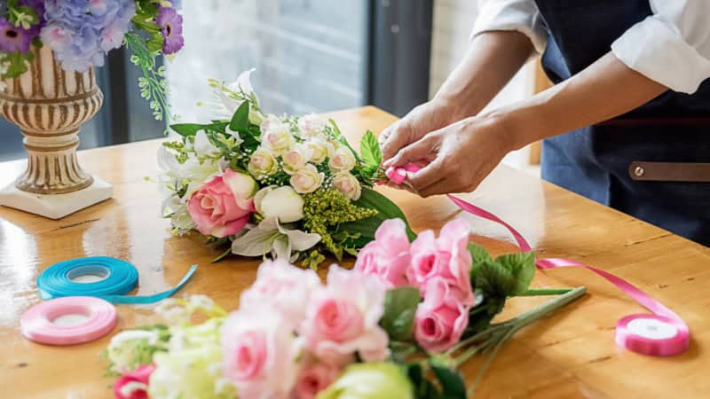 Eine Frau bindet einen Blumenstrauß auf einem Blumenstraußständer und dekoriert ihn mit einem Dekoband.