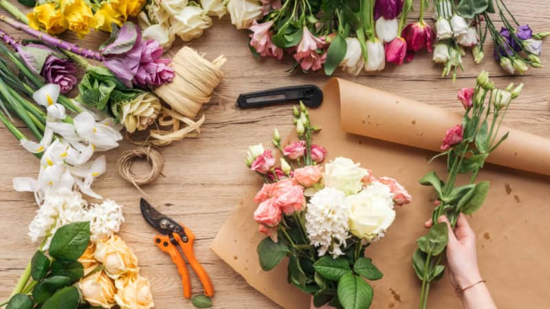 Eine Frau bindet einen Blumenstrauß auf einem Blumenstraußständer. Blumen und Werkzeuge liegen auf dem Tisch.