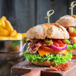 2 BBQ Bacon Burgers auf einem Schneidebrett, Kartoffelecken im Hintergrund.