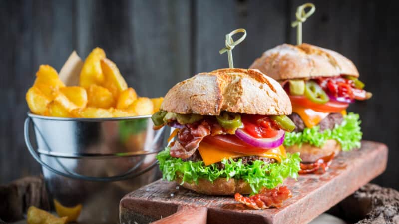 2 BBQ Bacon Burgers auf einem Schneidebrett, Kartoffelecken im Hintergrund.