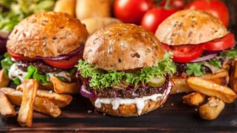 3 Classic Beef Burgers auf einem Schneidebrett, Gemüse im Hintergrund.