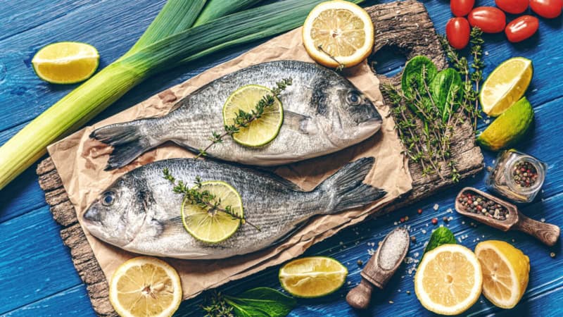Fischgericht auf einem Schneidebrett, Gemüse und Zitronen im Hintergrund.