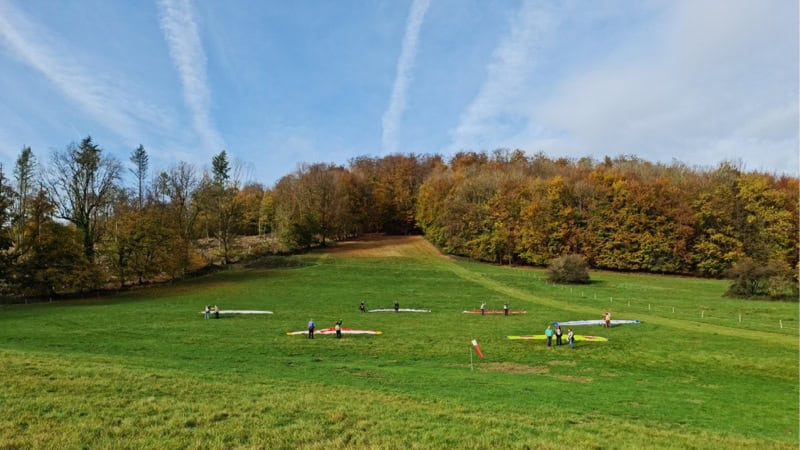 Die Schirme und Drachen liegen auf dem Hang auf der Wiese. Blauer Himmel und Bäume