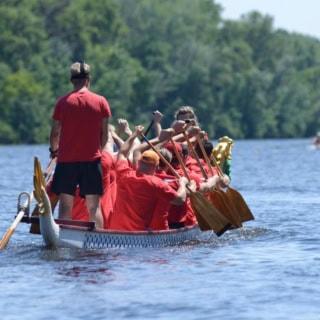 Ein Drachenboot mit Menschen die rote Shirts anhaben, wird von hinten fotografiert, wie es über einen Fluss fährt.