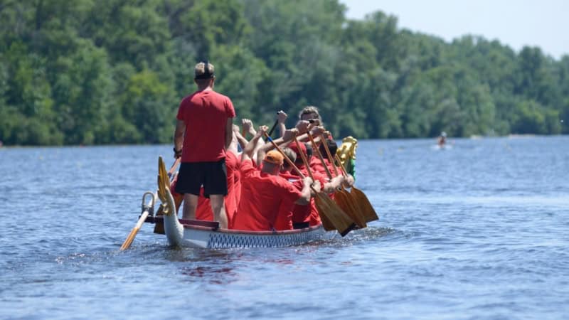 Ein Drachenboot mit Menschen die rote Shirts anhaben, wird von hinten fotografiert, wie es über einen Fluss fährt.