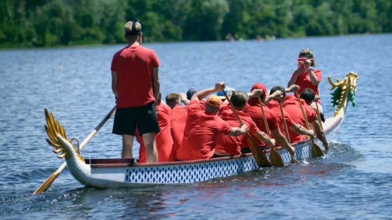 Ein Drachenboot mit Menschen in roten Shirts, wird von der Seite fotografiert, wie es über einen Fluss fährt.