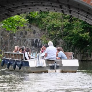 Motorboot mit vier Passagieren fährt unter einer Brücke auf dem Leipziger Kanal, dichtes Grün an den Ufern.