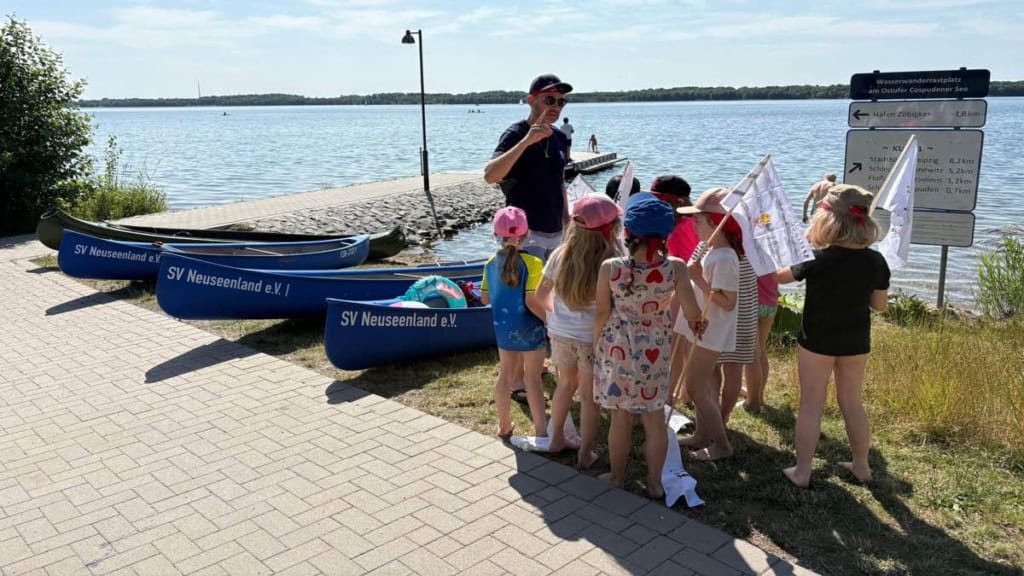 Eine Gruppe von Kindern mit Piratenflaggen in der Hand steht vor Kajaks die am Ufer des Sees liegen