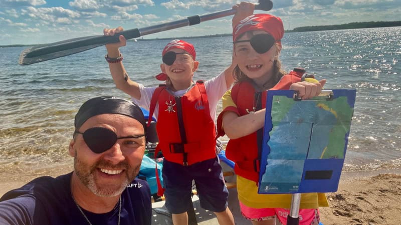 Ein Mann und 2 Kinder die als Piraten verkleidet sind stehen am Ufer eines Sees und ein Kind hält eine Schatzkarte