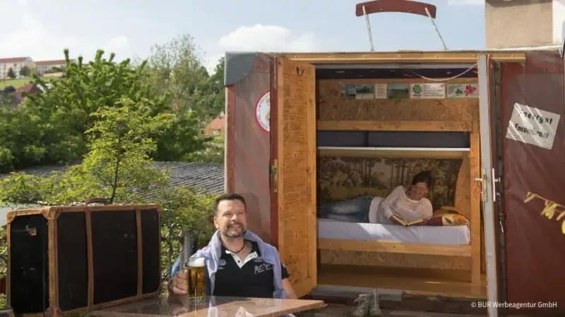 Frau liegt im Kofferhotel und Mann sitzt vor dem Hotel am Tisch und trinkt ein Bier