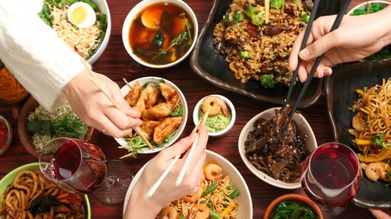 Verschiedene asiatische Gerichte stehen auf einem Tisch von dem sich Menschen mit Stäbchen bedienen.