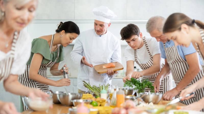 Ein Koch der mit Kochkurs Teilnehmern um einen Tisch steht und diesen etwas erklärt.