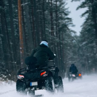 Zwei Personen fahren auf Quads im Winter durch einen Wald.