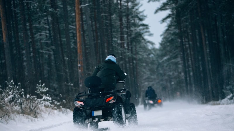 Zwei Personen fahren auf Quads im Winter durch einen Wald.