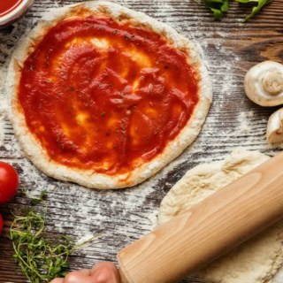 Ein Pizza Teig wird in Mehl ausgerollt. Ein Pizza Teig der mit Tomatensoße bestrichen wurde liegt daneben.