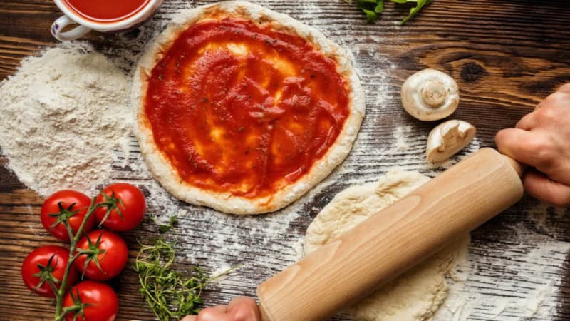 Ein Pizza Teig wird in Mehl ausgerollt. Ein Pizza Teig der mit Tomatensoße bestrichen wurde liegt daneben.
