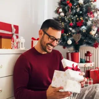 Ein Mann freut sich beim Öffnen eines Geschenks am Weihnachtsbaum.