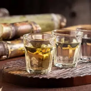 Drei mit Chachaca gefüllte Shotgläser stehen auf einem Brett aufgereiht auf einem Tisch. Im Hintergrund liegt Zuckerrohr auf dem Tisch.