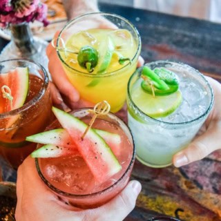 Vier unterschiedliche und bunte Cocktails werden über einem Tisch angestoßen.