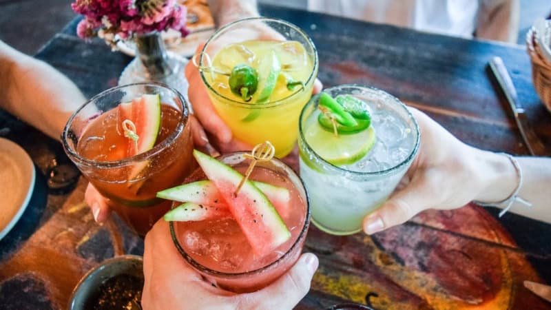 Vier unterschiedliche und bunte Cocktails werden über einem Tisch angestoßen.