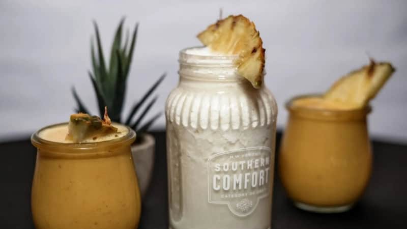 Drei verschiedene Gläser mit unterschiedlichen Cocktails und Pina-Colada stehen auf einem schwarzen Tisch. In jedem Cocktail steckt ein Stück Ananas.