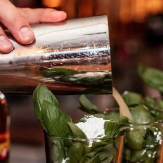 Eine Person schüttet einen Drink aus einem Cocktailshaker in vier vorbereitete Cocktail Gläser.