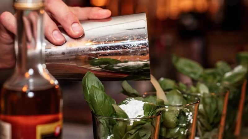 Eine Person schüttet einen Drink aus einem Cocktailshaker in vier vorbereitete Cocktail Gläser.