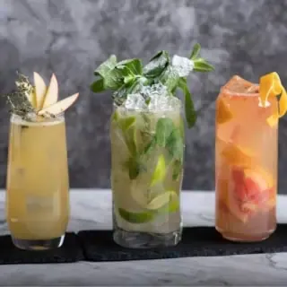 Fünf verschiedene und bunte Cocktails in unterschiedlicher Größe stehen aufgereiht auf einem Untersetzer vor einem dunkelgrauen Hintergrund.