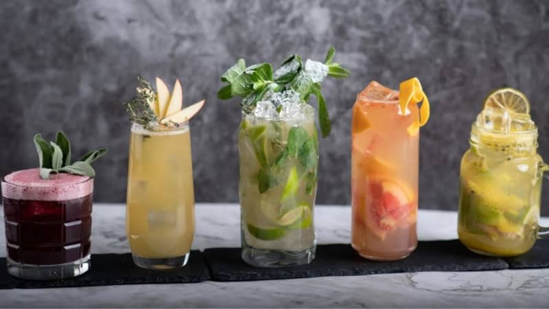 Fünf verschiedene und bunte Cocktails in unterschiedlicher Größe stehen aufgereiht auf einem Untersetzer vor einem dunkelgrauen Hintergrund.