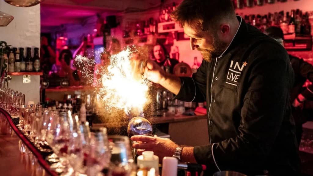 Cocktailkurs-in-Frankfurt-Tischfeuerwerk Ein Barkeeper steht hinter dem Tresen einer Bar und bereitet Cocktails vor. Daneben explodiert ein Tischfeuerwerk.