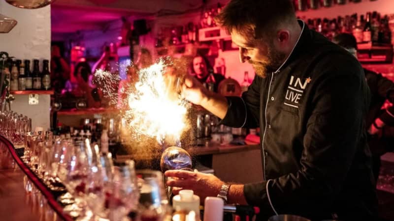 Ein Barkeeper steht hinter dem Tresen einer Bar und bereitet Cocktails vor. Daneben explodiert ein Tischfeuerwerk.