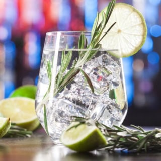 Nahaufnahme von einem Glas mit Gin und Gurken in einer Bar. Daneben liegen Limetten und Kräuter.